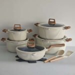 Cream ribbed ceramic-coated cookware set with glass lids and wood-style handles, including pots, pan, and utensils.